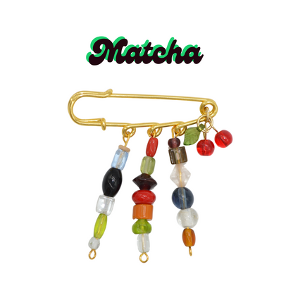 Gold brooch with colorful beads on a white background, featuring the brand 'Matcha'.